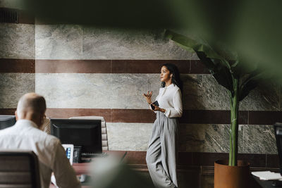 Confident female financial advisor talking through in-ear headphones leaning on wall at office