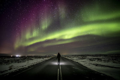Silhouette of unrecognizable traveler standing on asphalt roadway enjoying view of green polar lights glowing in night starry sky in iceland