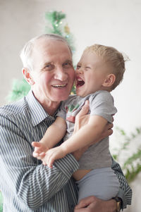 Grandfather holds his grandson in his arms and both laugh against the background of  christmas tree
