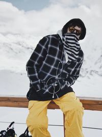 Portrait of man wearing sunglasses and warm clothing while sitting on fence during winter