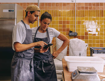Young couple of small business owners using mobile phone and making order to supplier while working together in bakery