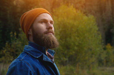 Young attractive bearded man in a red hat and blue jacket stands against the backdrop of a dense