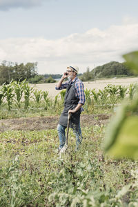 Gardener talking on mobile phone while standing with shovel at farm against sky