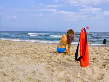 Rear view of woman in bikini kneeling on sand at beach against sky