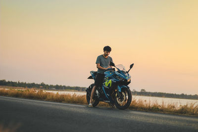 Man sitting on motorcycle at road against sky during sunset