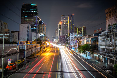 Light trails on city street by buildings against sky at night