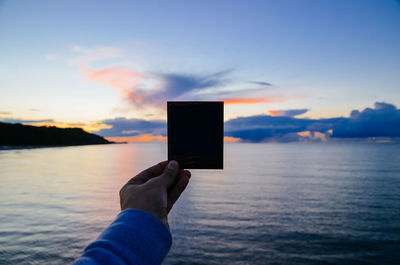 Cropped image of person holding instant film over sea against sky