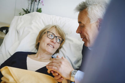 Smiling woman looking at senior partner while lying on bed in hospital ward