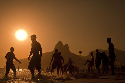 Silhouette friends playing soccer at beach against orange sky during sunset