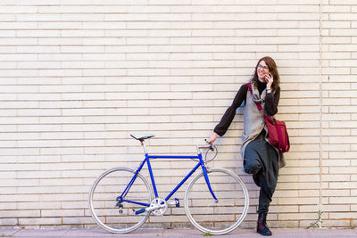 Smiling young woman with bike leaning on wall while using mobile