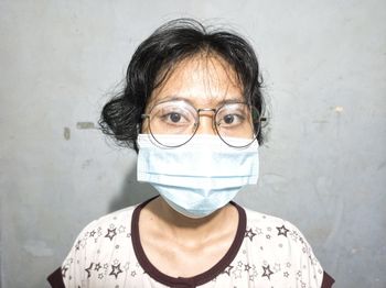 Portrait of woman wearing surgical mask and eyeglasses leaning against a wall