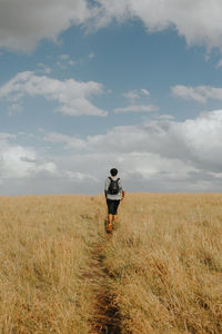 Rear view of mid adult man with backpack walking on grassy field against cloudy sky during sunny day