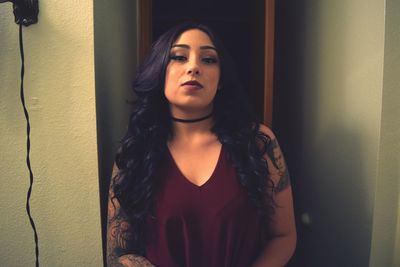 Portrait of tattooed young woman with curly hair standing by wall at home