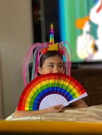 Cute girl covering mouth by multicolored hand paper fan while sitting on table at home