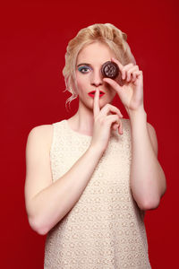 Portrait of woman with finger on lips holding chocolate chip cookie in front of eye while standing against red background