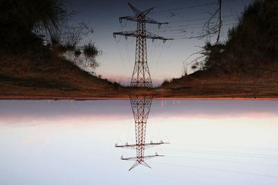Electricity pylon by lake against sky during sunset
