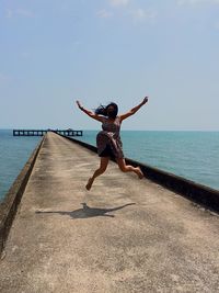 Woman with arms outstretched jumping on pier by sea against sky during sunny day