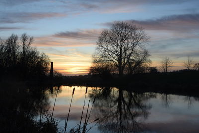 Silhouette bare trees by lake against sky during sunset