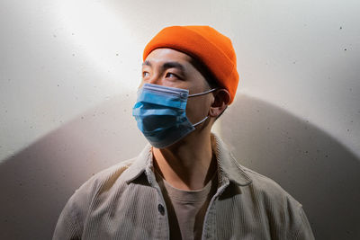 Concentrated asian male in casual wear and protective face mask standing calmly in blurred gray room and looking away