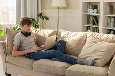 A teenager boy sits on a beige sofa, in room, wearing black headphones around , looks into a laptop.