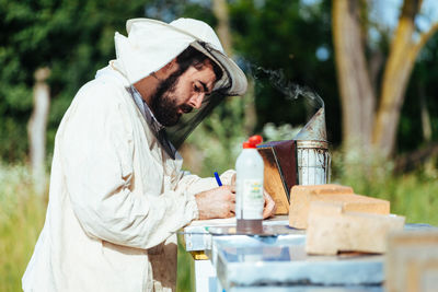 Side view of beekeeper writing in book on beehive 