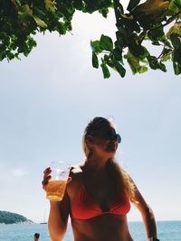 Woman in bikini drinking beer at beach against sky