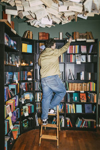 Rear view of young man choosing books while standing on ladder in bookstore