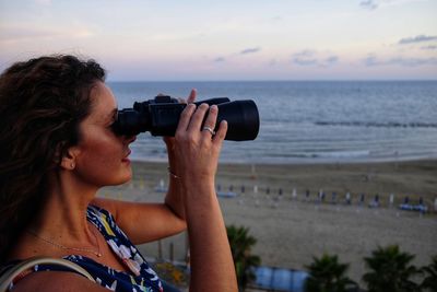 Woman looking through binoculars standing against beach during sunset