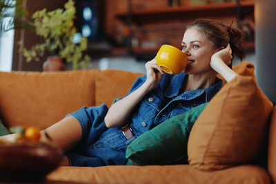 Young woman drinking drink while sitting on sofa at home