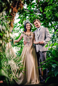 Portrait of smiling bride and groom holding hands while standing amidst plants