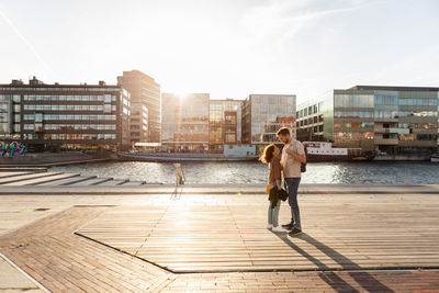 Full length of loving couple standing on promenade by river in city during summer