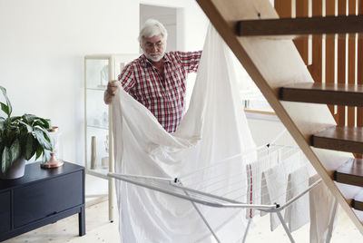 Senior man folding white sheet for drying on rack in living room