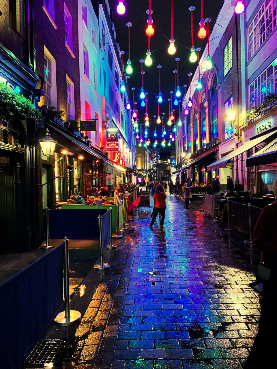 PEOPLE WALKING ON ILLUMINATED STREET AMIDST BUILDINGS IN CITY