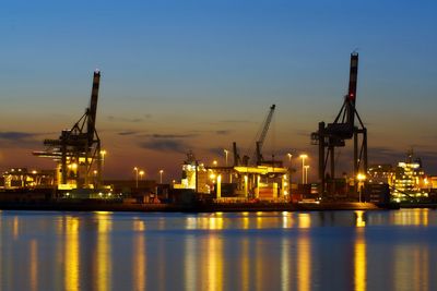 Illuminated commercial dock by river against sky during sunset