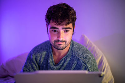 Man using digital tablet sitting on bed against wall at home