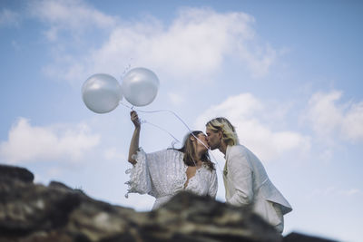 Romantic bride holding white helium balloons kissing groom against sky