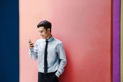 Smiling young businessman using phone while standing against wall
