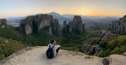 Rear view of man sitting on rock against sky during sunset