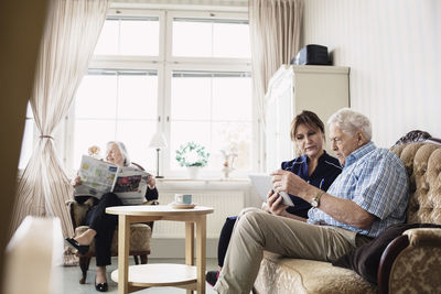 Senior man and caretaker using digital tablet while woman reading newspaper in nursing home