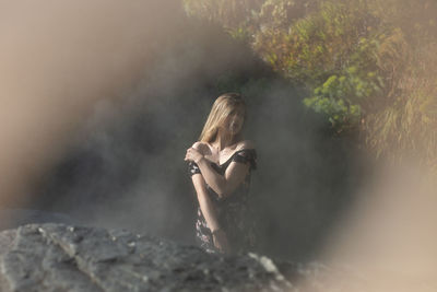 Beautiful young woman looking away while sitting in rock against plants