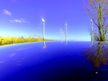 Illuminated street light on field by trees against blue sky