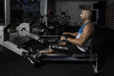 Side view of determined disabled man exercising on rowing machine at gym