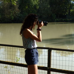 Young woman photographing while standing by railing against lake