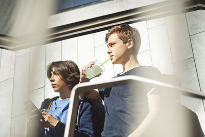 Boy looking away while drinking juice with friend holding smart phone outdoors