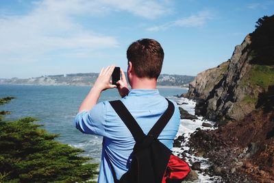 Rear view of man with backpack photographing with mobile phone at beach