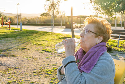 Elderly woman drinks water from the bottle after exercising in a park at sunset