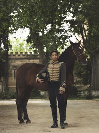 Full length of adult black female in elegant outfit standing with brown horse near green trees and castle fence in daytime in yard