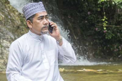 Thoughtful man talking on phone while sitting against waterfall