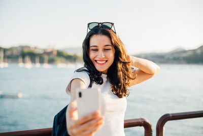 Portrait of smiling young woman holding camera while standing against sea