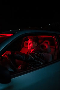 Side view of confident young male millennial sitting in modern car with red neon illumination at night and looking away